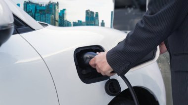İlerici işadamı elektrikli aracını şarj kablosu ile şarj ediyor. Şarj istasyonundan şehrin arka planına bakıyor. Modern temiz enerji kampanyası konsepti için..