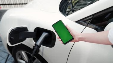 Taklit uzay için şarj istasyonuna bağlı elektrikli aracın yeşil ekran enerji durumunu gösteren bir model telefon. Temiz çevre için ilerici bir kavram. EV, yeşil yenilenebilir enerjiden güç alıyor.