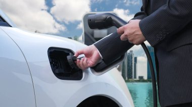 İlerici işadamı elektrikli aracını şarj kablosu ile şarj ediyor. Şarj istasyonundan şehrin arka planına bakıyor. Modern temiz enerji kampanyası konsepti için..