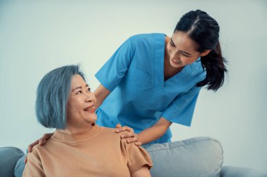 A caregiver rest her hands on the shoulders of a contented senior patient while she sitting on the sofa at home.