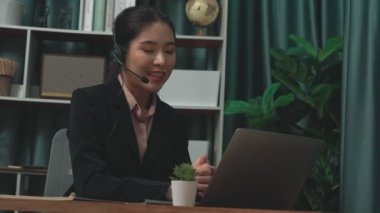 Kulaklık ve mikrofon takan iş kadını masasında çalışıyor ve dizüstü bilgisayar kullanıyor. Uzaktan kumandalı online toplantı için dizüstü bilgisayar kullanan hevesli kadın çalışanlar, ofiste iş videosu konferansı.