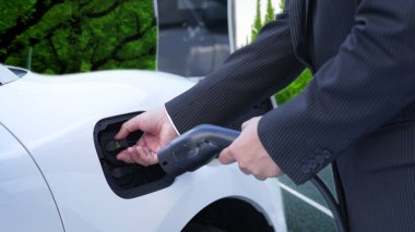 İlerici işadamı elektrikli aracını şarj kablosu ile şarj ediyor. Şarj istasyonundan şehrin arka planına bakıyor. Modern temiz enerji kampanyası konsepti için..