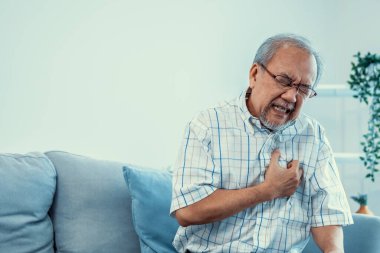 An agonizing senior man suffering from chest pain or heart attack alone in his living room. Serious health problem and feeling unwell concept for seniors.