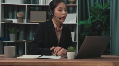 Kulaklık ve mikrofon takan iş kadını masasında çalışıyor ve dizüstü bilgisayar kullanıyor. Uzaktan kumandalı online toplantı için dizüstü bilgisayar kullanan hevesli kadın çalışanlar, ofiste iş videosu konferansı.