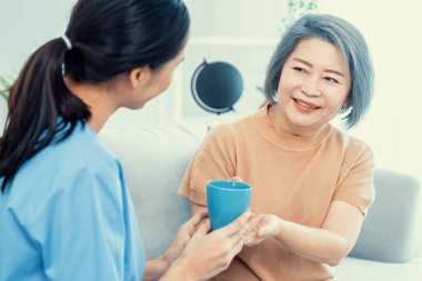 Female care taker serving her contented senior patient with a cup of coffee at home, smiling to each other. Medical care for pensioners, Home health care service.