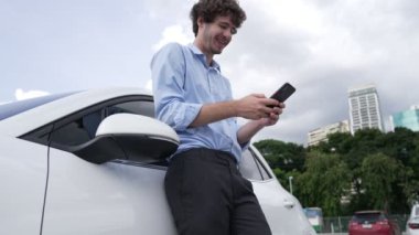 Takım elbiseli, hırslı bir işadamı elektrik kablosu ve yenilenebilir enerji enerjili bir elektrikli araçla şarj istasyonunda dururken elektrikli aracına yaslanıyor..