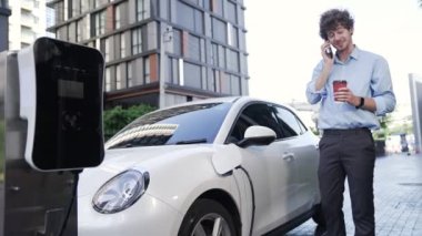 Takım elbiseli, hırslı bir işadamı elektrik kablosu ve yenilenebilir enerji enerjili bir elektrikli araçla şarj istasyonunda dururken elektrikli aracına yaslanıyor..