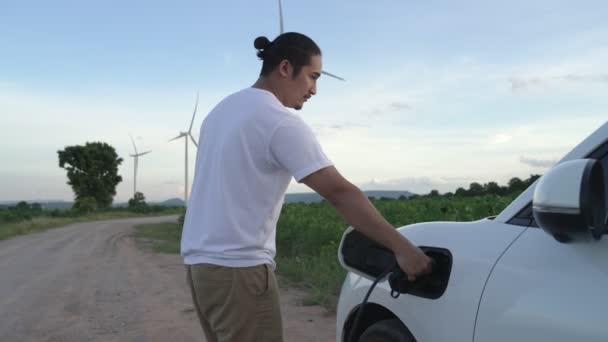 将来の持続可能なエネルギーの概念として風力タービンと緑のフィールド上の充電ステーションからのエネルギーを充電Ev車 彼の電気自動車と進歩的な男 発電機付き電気自動車 — ストック動画