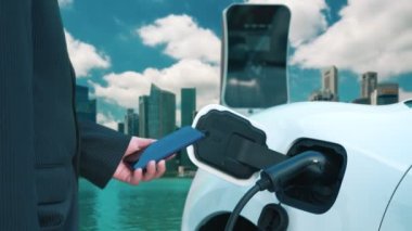 İlerici işadamı elektrikli aracı şarj kablosu ile şarj etme istasyonu ve temiz enerji kampanyası için şehir arka planına sahip akıllı telefondan enerji durumunu kontrol etme.
