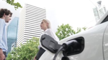İlerici işadamı ve iş kadını, şehir merkezinde dolaşmadan önce şarj cihazını şarj istasyonundan elektrikli arabaya takıyor. Çevre dostu şarj edilebilir araba temiz enerjiyle çalışıyor..