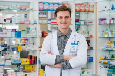 Portrait of a qualified, friendly male pharmacist wearing a white coat, crossing his arms, and looking at the camera, with a shelf of various medicine boxes in background at drugstore or pharmacy. clipart