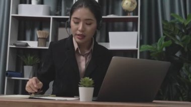 Kulaklık ve mikrofon takan iş kadını masasında çalışıyor ve dizüstü bilgisayar kullanıyor. Uzaktan kumandalı online toplantı için dizüstü bilgisayar kullanan hevesli kadın çalışanlar, ofiste iş videosu konferansı.