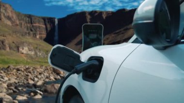 Elektrikli araba, doğal ortam, şelale ve akış arkaplanındaki şarj istasyonundan enerji yüklüyor. Yenilenebilir enerji kaynaklı EV otomobillerin enerji sürdürülebilirliği kavramı.