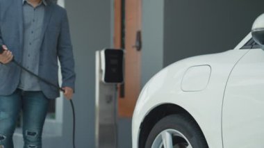 İlerici adam evindeki elektrikli aracın aküsüne emisyonsuz güç bağlantısı kuruyor. Elektrikli araç elektrik istasyonundan EV araba aküsüne kabloyla şarj oluyor.
