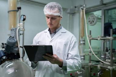 Eczacı bilim adamı bir CBD yağ çıkarıcısından bilgi almak için bir pano ve kalem kullanıyor ve tıbbi esrar ürünleri üretmek için bilimsel bir makine kullanıyor..