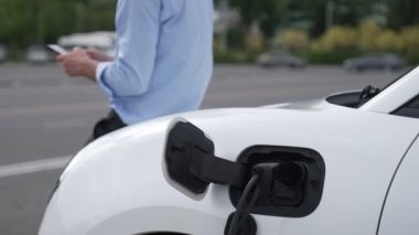 Takım elbiseli, hırslı bir işadamı elektrik kablosu ve yenilenebilir enerji enerjili bir elektrikli araçla şarj istasyonunda dururken elektrikli aracına yaslanıyor..