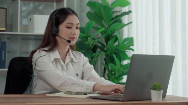 Asian customer support operator wearing headset and microphone working at her desk with laptop. Enthusiastic female employee provide customer service, supportive call center agent helping customers. clipart