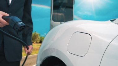 İlerici işadamı elektrikli arabayı tepe üzerindeki yolculuğunda şarj istasyonuyla dolduruyor. Temiz çevre için yenilenebilir enerji ile çalışan EV arabası. EV otomobilinden enerji sürdürülebilirliği kavramı.