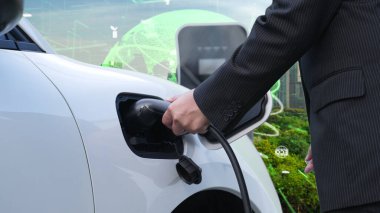 İlerleyen yeşil şehir ESG sembolü, elektrikli araçla birlikte. İş adamı EV arabalarını şarj etmek için şarj aletini şarj ediyor. Şehirdeki şarj istasyonunda ekolojik ve yeşil semboller var..