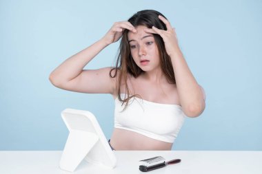 Young charming teenager girl worry about her face pimples on mirror for skincare and acne problem concept. Stress young girl squeezing pimples and pointing fingers on acne on her face.