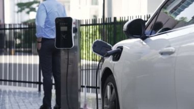 Takım elbiseli, hırslı bir işadamı elektrik kablosu ve yenilenebilir enerji enerjili bir elektrikli araçla şarj istasyonunda dururken elektrikli aracına yaslanıyor..