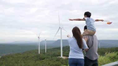 İlerici mutlu aile kavramı rüzgâr türbini çiftliğinde eğleniyor. Rüzgar türbini jeneratörüyle gelen elektrik jeneratörü kırsal bölgede tepe ve ufukta dağ var..
