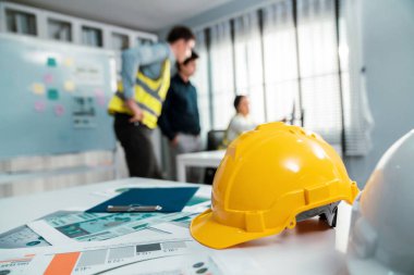 Concept of engineering equipments of protective helmet for competent engineer on piles of paper works. Blurred image of engineers and employer working together at the desk.