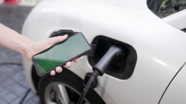Taklit uzay için şarj istasyonuna bağlı elektrikli aracın yeşil ekran enerji durumunu gösteren bir model telefon. Temiz çevre için ilerici bir kavram. EV, yeşil yenilenebilir enerjiden güç alıyor.
