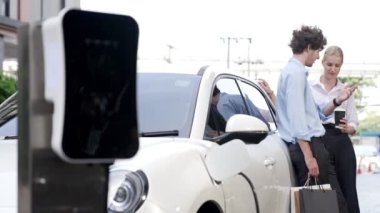 İlerici iş adamı ve iş kadını şehir merkezinde dolaşmadan önce şarj istasyonuna bağlı elektrikli arabaya yaslanıyor. Çevre dostu şarj edilebilir araba temiz enerjiyle çalışıyor..
