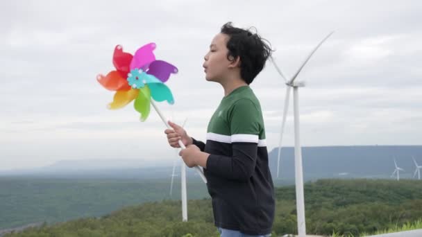 丘の上の風力タービン農場 緑のフィールドで風力タービンのおもちゃで遊んで進歩的な若いアジアの少年 再生可能エネルギー発電からのグリーンエネルギー — ストック動画