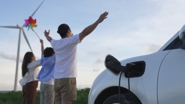 電気自動車で風力発電所での時間を楽しむ進歩的な幸せな家族の概念 風力発電機からの再生可能エネルギーを利用した充電ステーション用電気自動車 — ストック動画