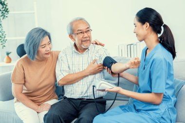 An elderly man having a blood pressure check by his personal caregiver with his wife sitting next to him in their home.