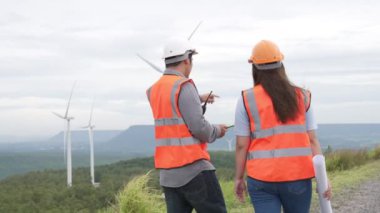 Erkek ve kadın mühendisler kırsal kesimdeki bir tepenin tepesinde ya da dağda rüzgar çiftliğinde çalışıyorlar. Yenilenebilir ve sürdürülebilir enerjinin gelecekteki üretimi için yenilikçi idealler.