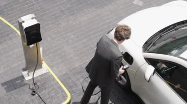 İlerici işadamı, şehir merkezinde dolaşmadan önce şarj cihazından elektrikli arabasına şarj prizi takıyor. Çevre dostu şarj edilebilir araba, sürdürülebilir ve temiz enerjiyle çalışıyor..