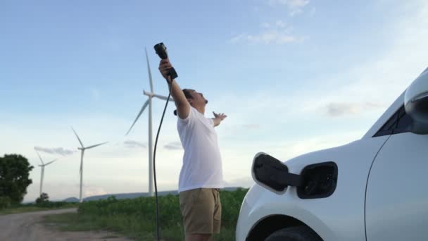 将来の持続可能なエネルギーの概念として風力タービンと緑のフィールド上の充電ステーションからのエネルギーを充電Ev車 彼の電気自動車と進歩的な男 発電機付き電気自動車 — ストック動画