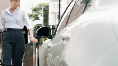 İlerici iş kadını şehir merkezinde dolaşmadan önce şarj cihazından elektrikli arabasına şarj prizi takıyor. Çevre dostu şarj edilebilir araba, sürdürülebilir ve temiz enerjiyle çalışıyor..