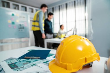 Concept of engineering equipments of protective helmet for competent engineer on piles of paper works. Blurred image of engineers and employer working together at the desk.