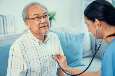 Caring young female doctor examining her contented senior patient with stethoscope in living room. Medical service for elderly, elderly sickness, declining health.