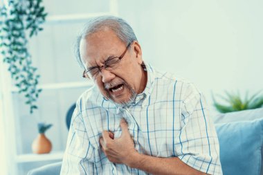 An agonizing senior man suffering from chest pain or heart attack alone in his living room. Serious health problem and feeling unwell concept for seniors.