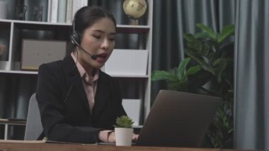 Kulaklık ve mikrofon takan iş kadını masasında çalışıyor ve dizüstü bilgisayar kullanıyor. Uzaktan kumandalı online toplantı için dizüstü bilgisayar kullanan hevesli kadın çalışanlar, ofiste iş videosu konferansı.