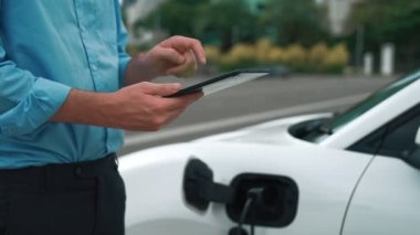 Takım elbiseli, hırslı bir işadamı elektrik kablosu ve yenilenebilir enerji enerjili bir elektrikli araçla şarj istasyonunda dururken elektrikli aracına yaslanıyor..
