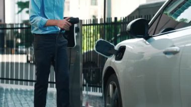 İlerici işadamı, şehir merkezinde dolaşmadan önce şarj cihazından elektrikli arabasına şarj prizi takıyor. Çevre dostu şarj edilebilir araba, sürdürülebilir ve temiz enerjiyle çalışıyor..
