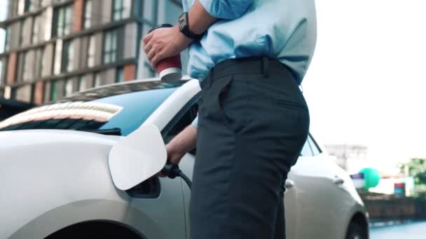 電源ケーブルプラグと再生可能エネルギー車と充電ステーションに立っている間 彼の電気自動車にもたれて進歩的な野心を持つスーツを身に着けているビジネスマン — ストック動画
