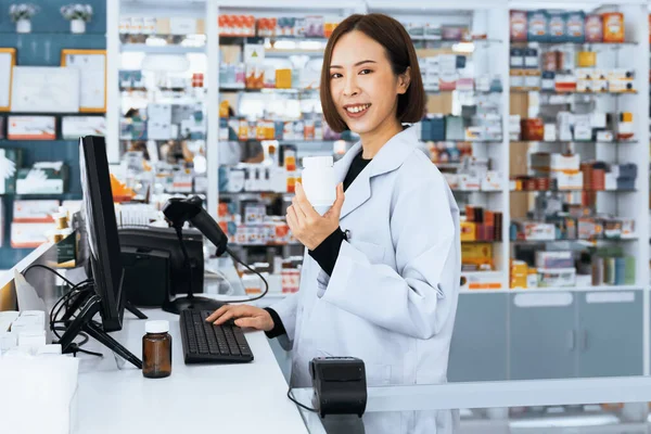 Portrait of young affable pharmacist and qualified pharmaceutical, medicine pill container or bottle mockup for copyspace at pharmacy. Druggist working with her diary job at drugstore, medicine shelf.