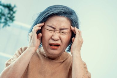 An agonizing senior woman with a headache, compress her temple with both hands at her contented living room. Senior health, nursing home, caretaker service.