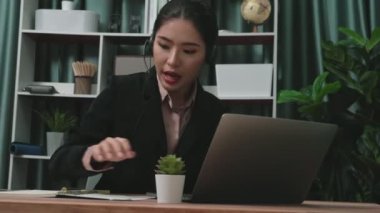 Kulaklık ve mikrofon takan iş kadını masasında çalışıyor ve dizüstü bilgisayar kullanıyor. Uzaktan kumandalı online toplantı için dizüstü bilgisayar kullanan hevesli kadın çalışanlar, ofiste iş videosu konferansı.