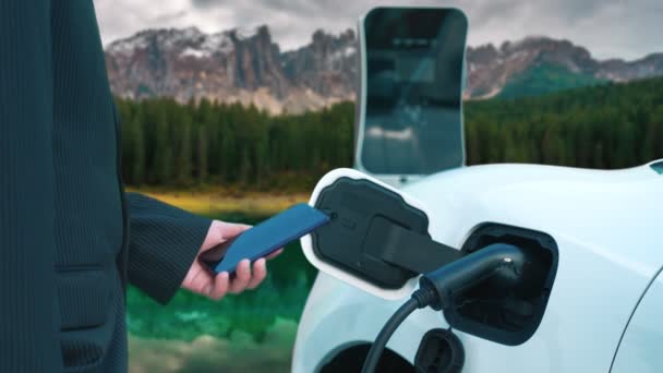 渐进式商人在有山地背景的充电站通过智能手机查看电动车电池的状况 现代技术与自然景观相结合 环保电动车 — 图库视频影像