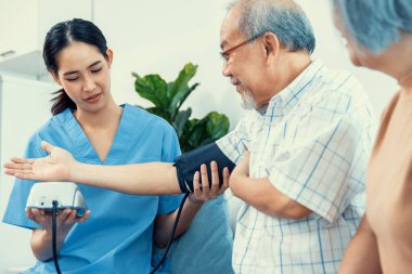 An elderly man having a blood pressure check by his personal caregiver with his wife sitting next to him in their home.