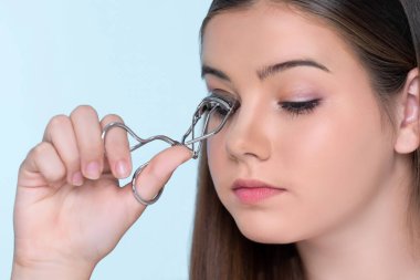 Closeup happy young charming brunette holding eyelash curler in her hand as makeup accessory. Beautiful girl applying eyelash curler in studio with isolated background for copyspace.