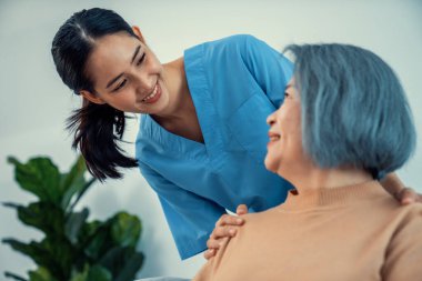 A caregiver rest her hands on the shoulders of a contented senior patient while she sitting on the sofa at home.